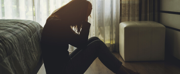 Sad woman sitting on floor against bed with hands on head.